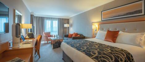 A hotel room with a large bed, a desk with a laptop, a chair, a sofa, and a TV.
