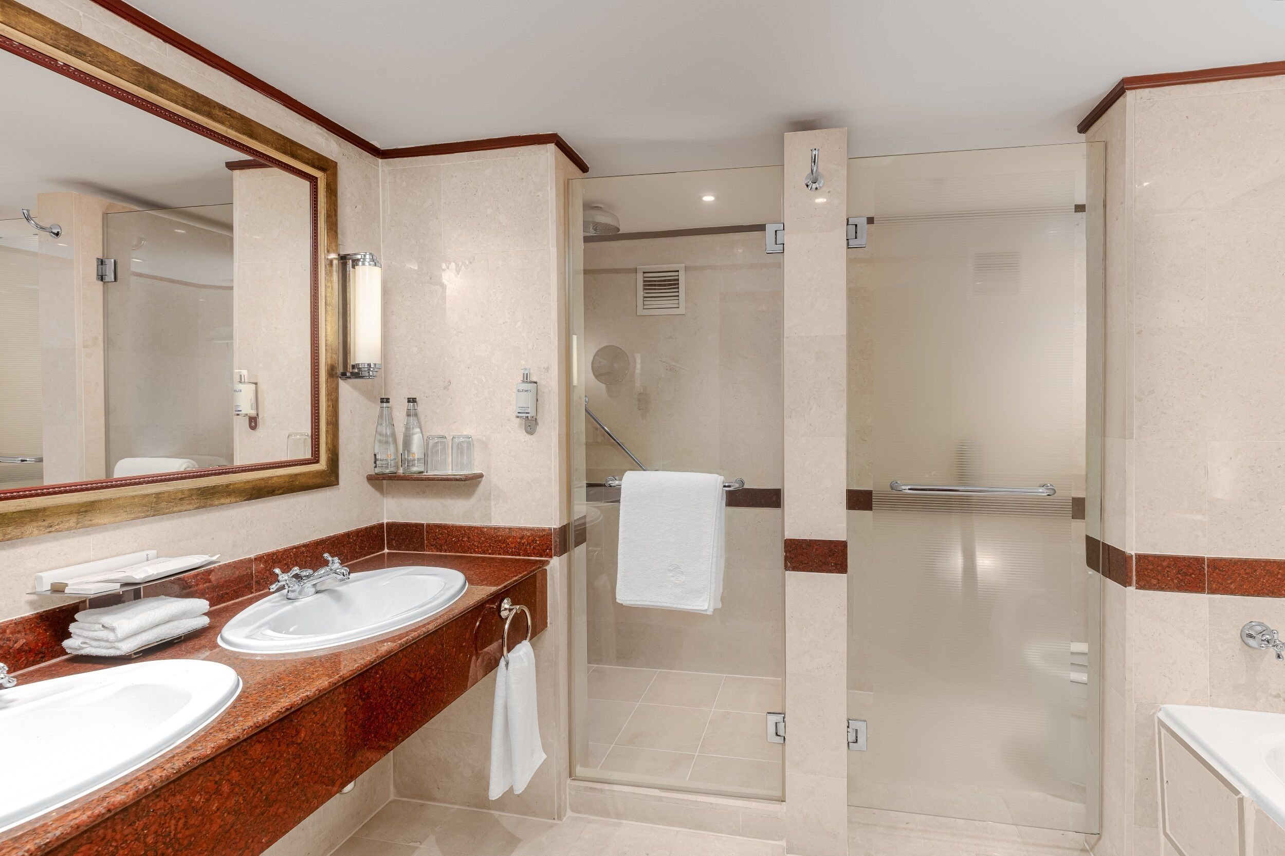 A hotel bathroom with two sinks, a large mirror, and a glass-enclosed shower area.