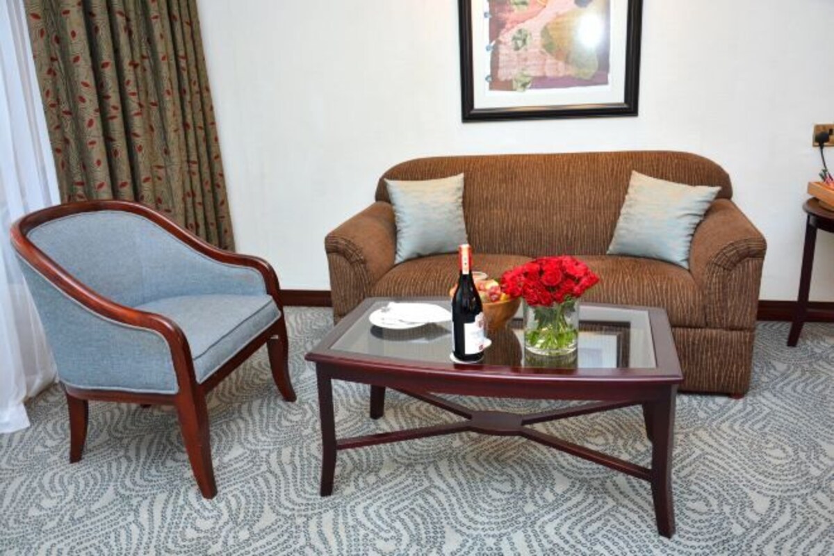 A living room with a brown sofa, a wooden armchair, and a glass coffee table with a bottle and flowers.
