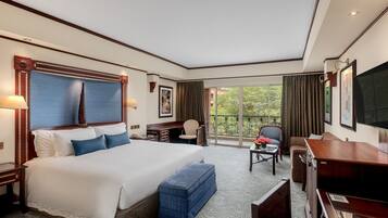 A hotel room with a large bed, a desk, a chair, a television, and a balcony with a view of trees.