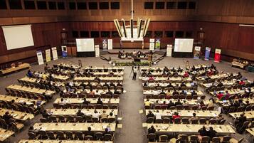 A large conference hall with multiple rows of tables and chairs, a podium at the front, and a large screen at the back.