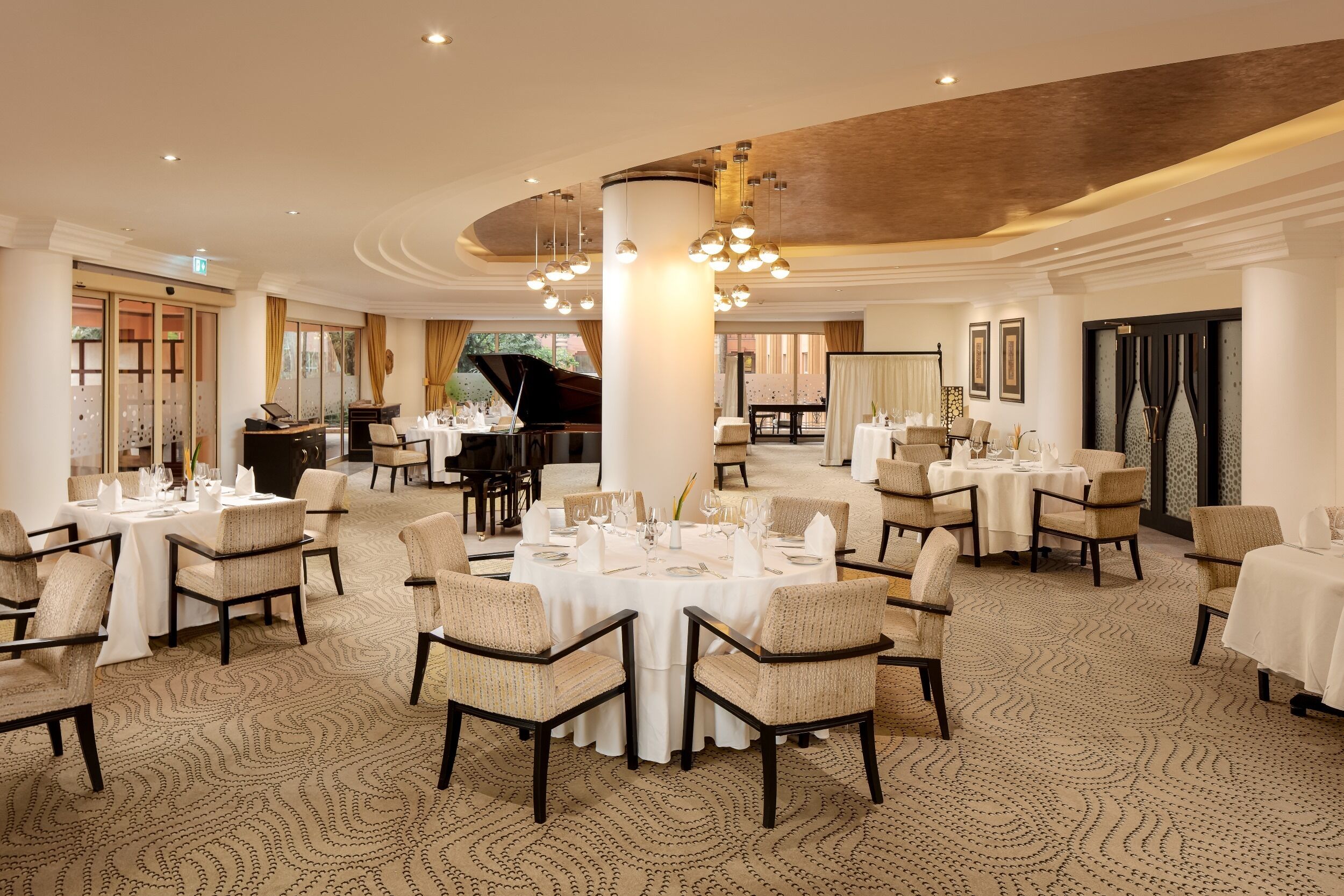 A spacious dining area with round tables and chairs, a grand piano, and a large chandelier.