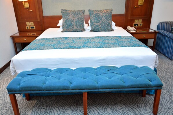 Habitación de hotel con una cama grande, dos almohadas iguales, una mesita de noche con un teléfono y un banco tapizado en azul.