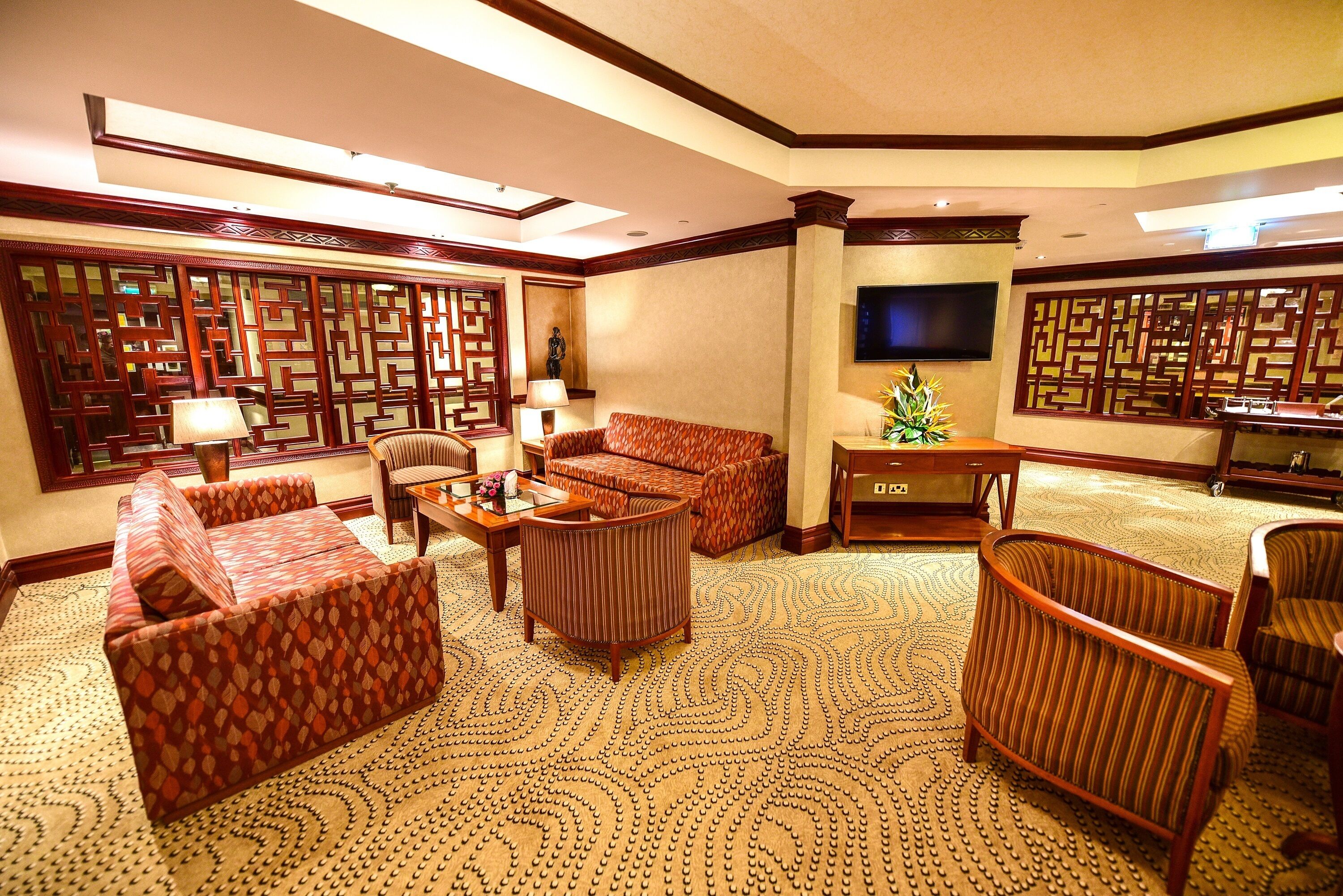 A spacious living room with a patterned carpet, wooden furniture, a flat-screen TV, and a decorative wall panel.