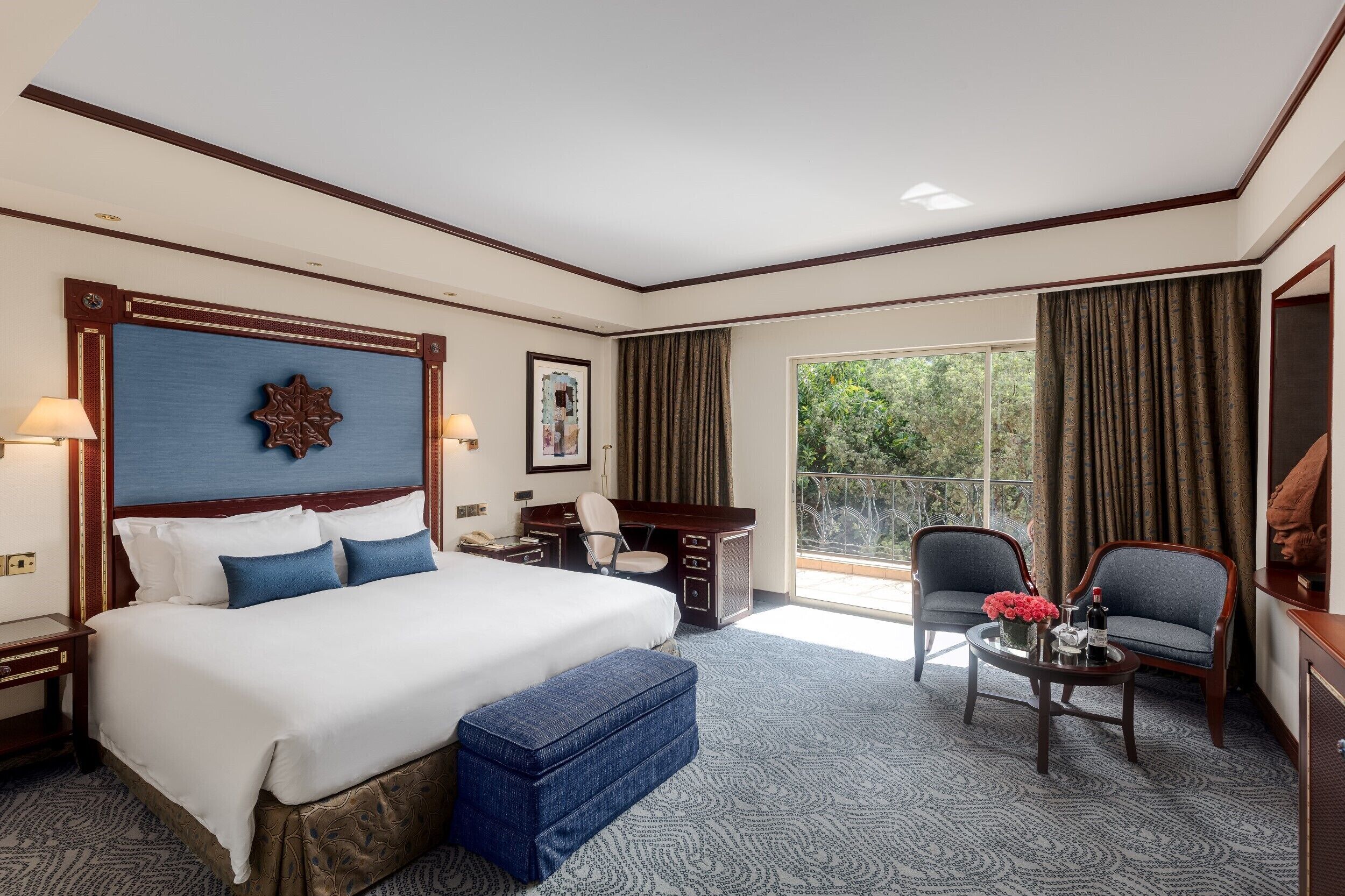 A hotel room with a large bed, a desk, a chair, a small table with a vase of flowers, and a view of greenery through the window.