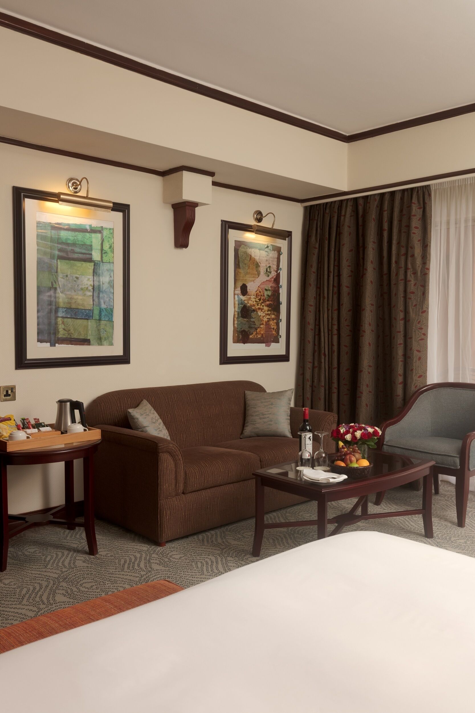 Une chambre d’hôtel avec un canapé marron, une petite table sur laquelle se trouve une machine à café, une chaise et un lit. Deux œuvres d’art encadrées sont accrochées au mur.