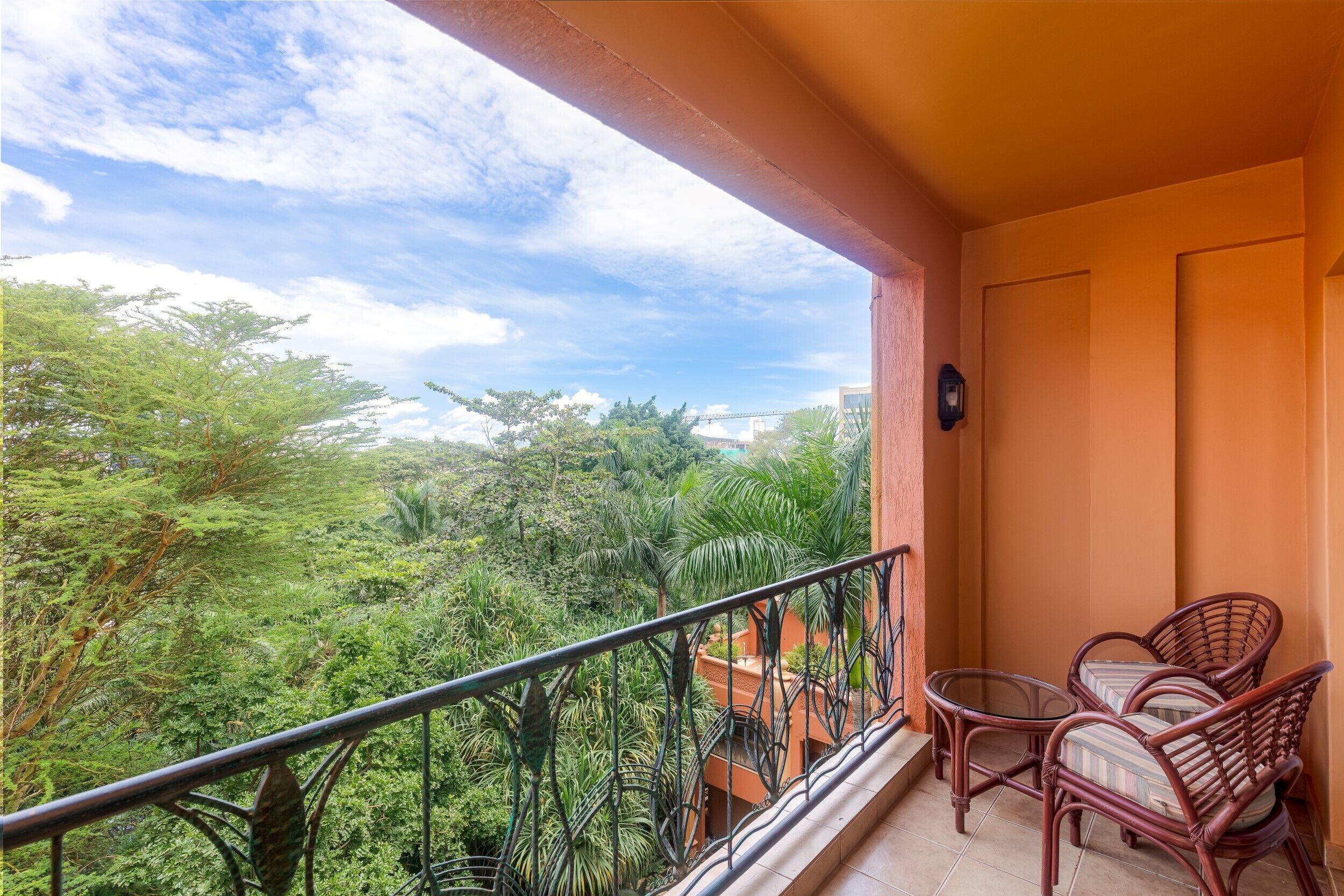 A balcony with a view of a lush green landscape and a distant bridge. There are two wicker chairs and a small table on the balcony.