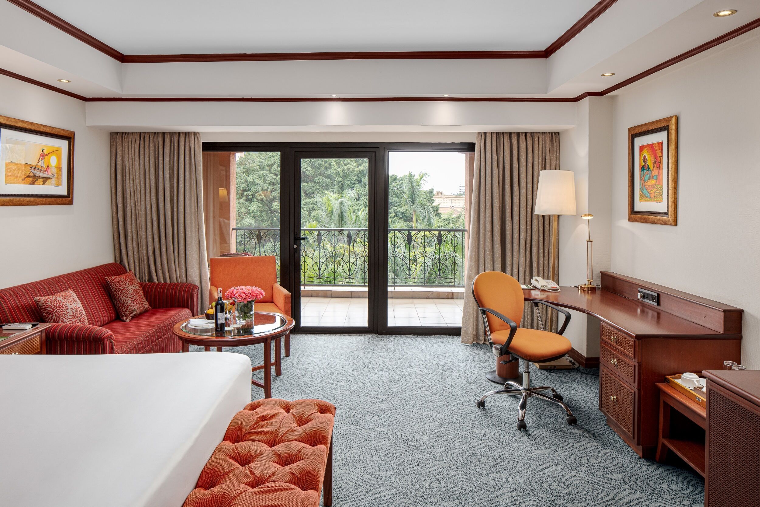 A hotel room with a bed, a sofa, a desk, a chair, and a balcony with a view of greenery.