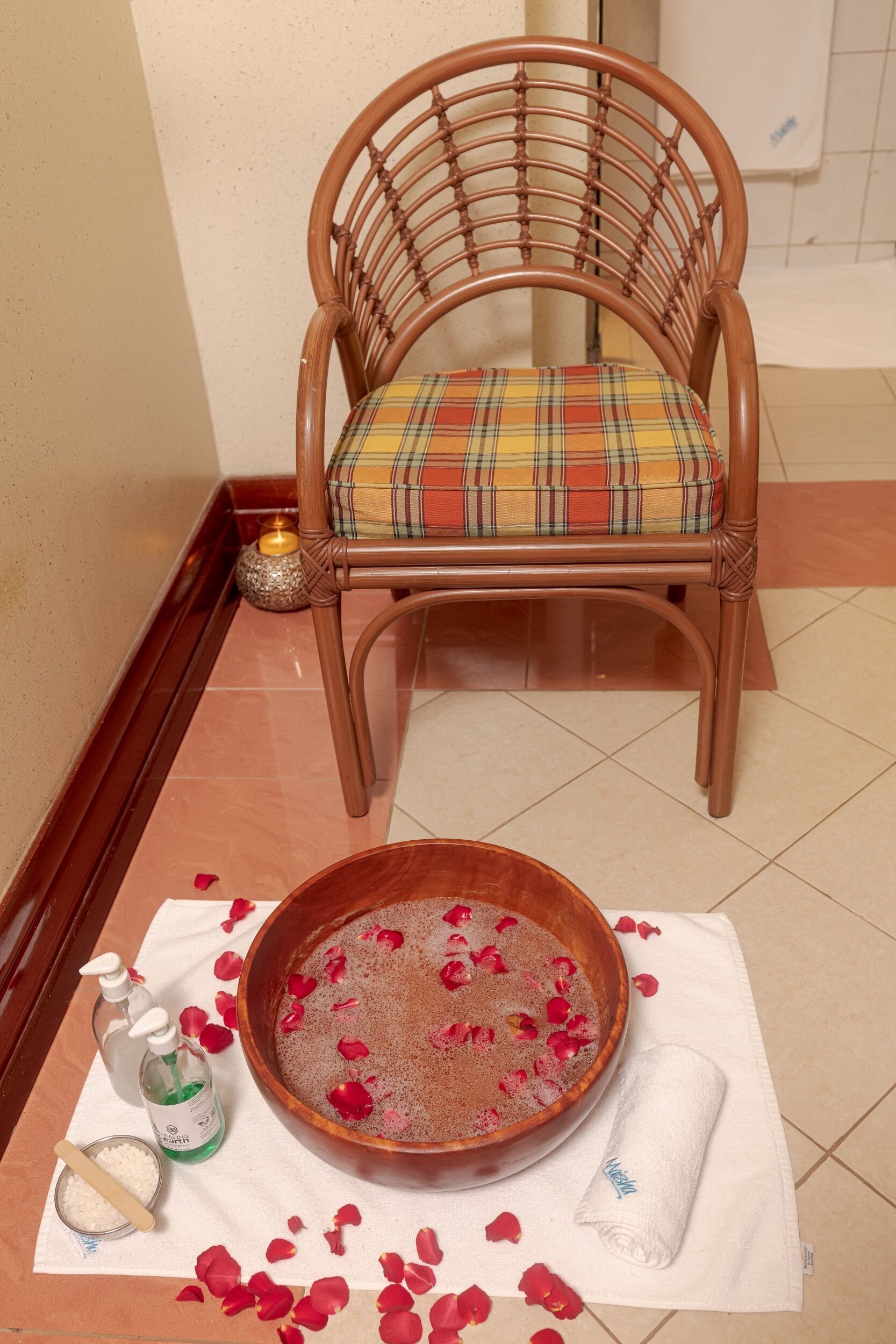 A wooden chair with a plaid cushion, a bowl with rose petals, and a bottle of soap on a white towel.