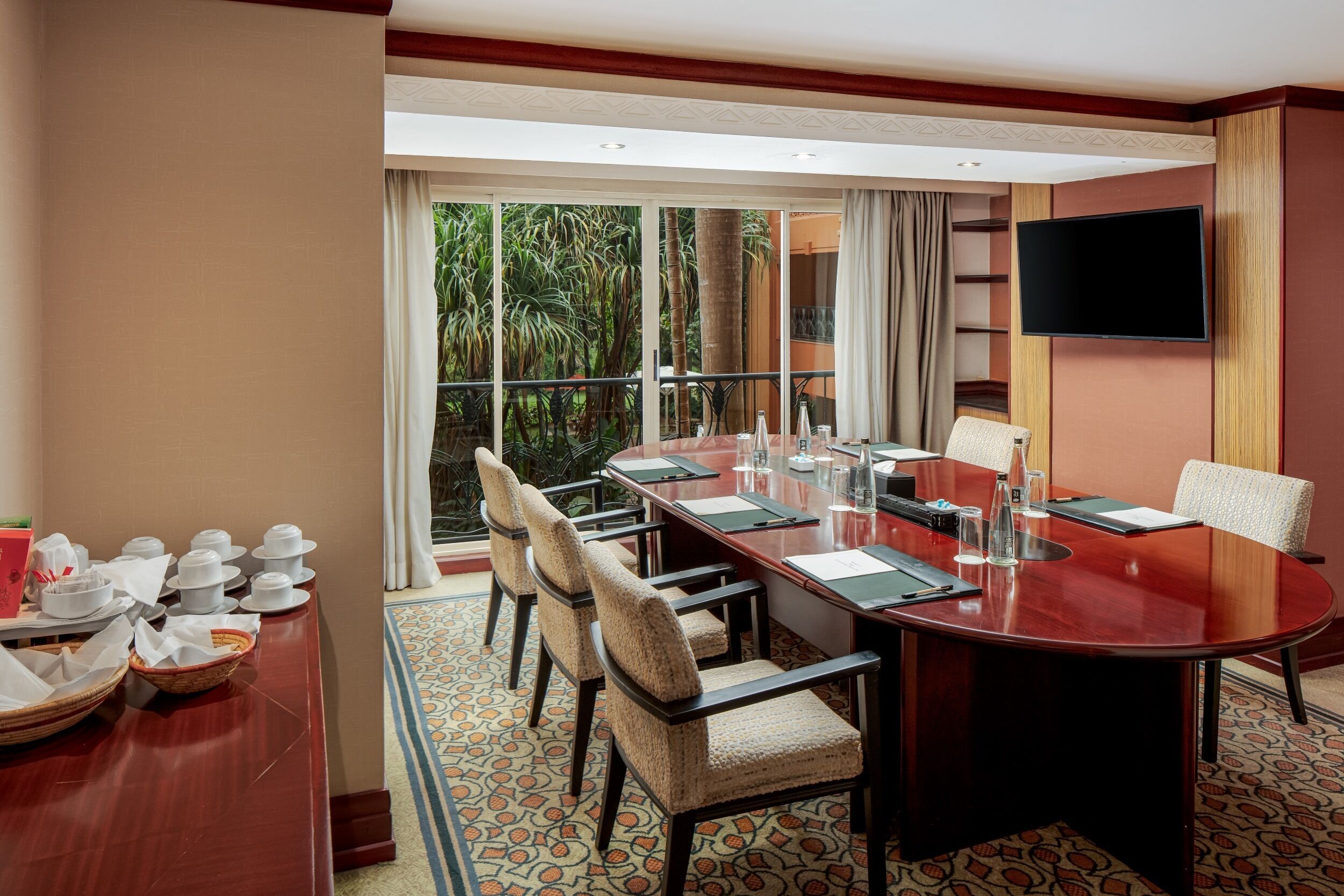 A hotel conference room with a long table, chairs, a TV, and a view of greenery outside.
