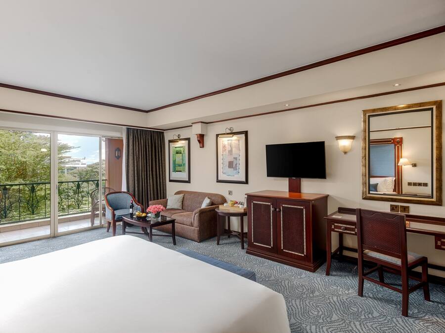 A hotel room with a large bed, a desk, a chair, a television, and a balcony with a view of trees.