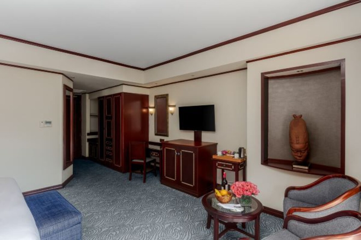 A hotel room with a bed, a desk with a television, a chair, a small table with a vase, and a wardrobe.