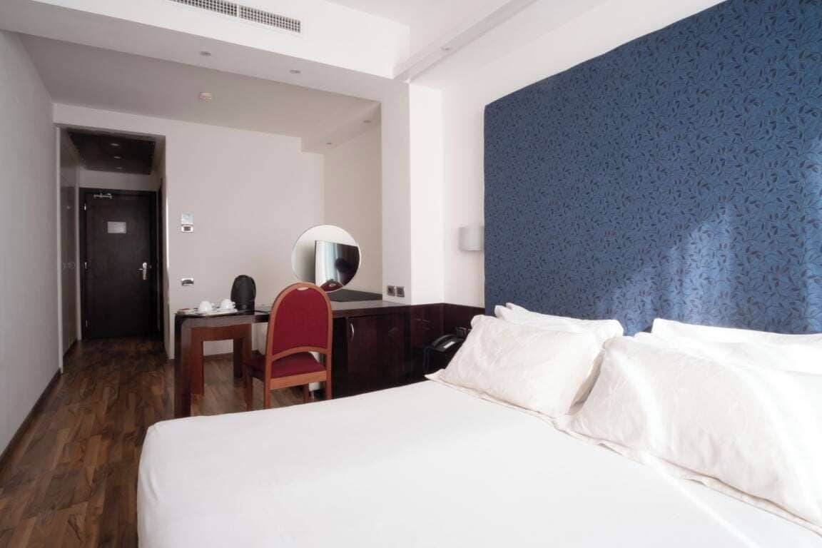 A hotel room with a bed, a desk, a chair, a mirror, and a blue textured wall.