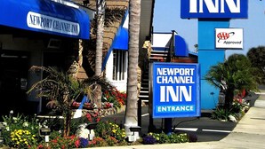 The exterior of the Newport Channel Inn with a clear entrance sign, palm trees, and a blue awning. - Newport Channel Inn - Near Huntington State Beach (Newport Beach)