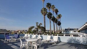 A rooftop terrace with white plastic chairs and tables, palm trees, and a clear blue sky. - Newport Channel Inn - Near Huntington State Beach (Newport Beach)