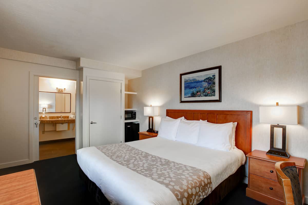 A hotel room with a large bed, a wooden headboard, a nightstand with a lamp, a dresser, and a bathroom visible through an open door.