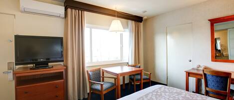 A hotel room with a bed, a desk, a chair, a television, and an air conditioning unit.