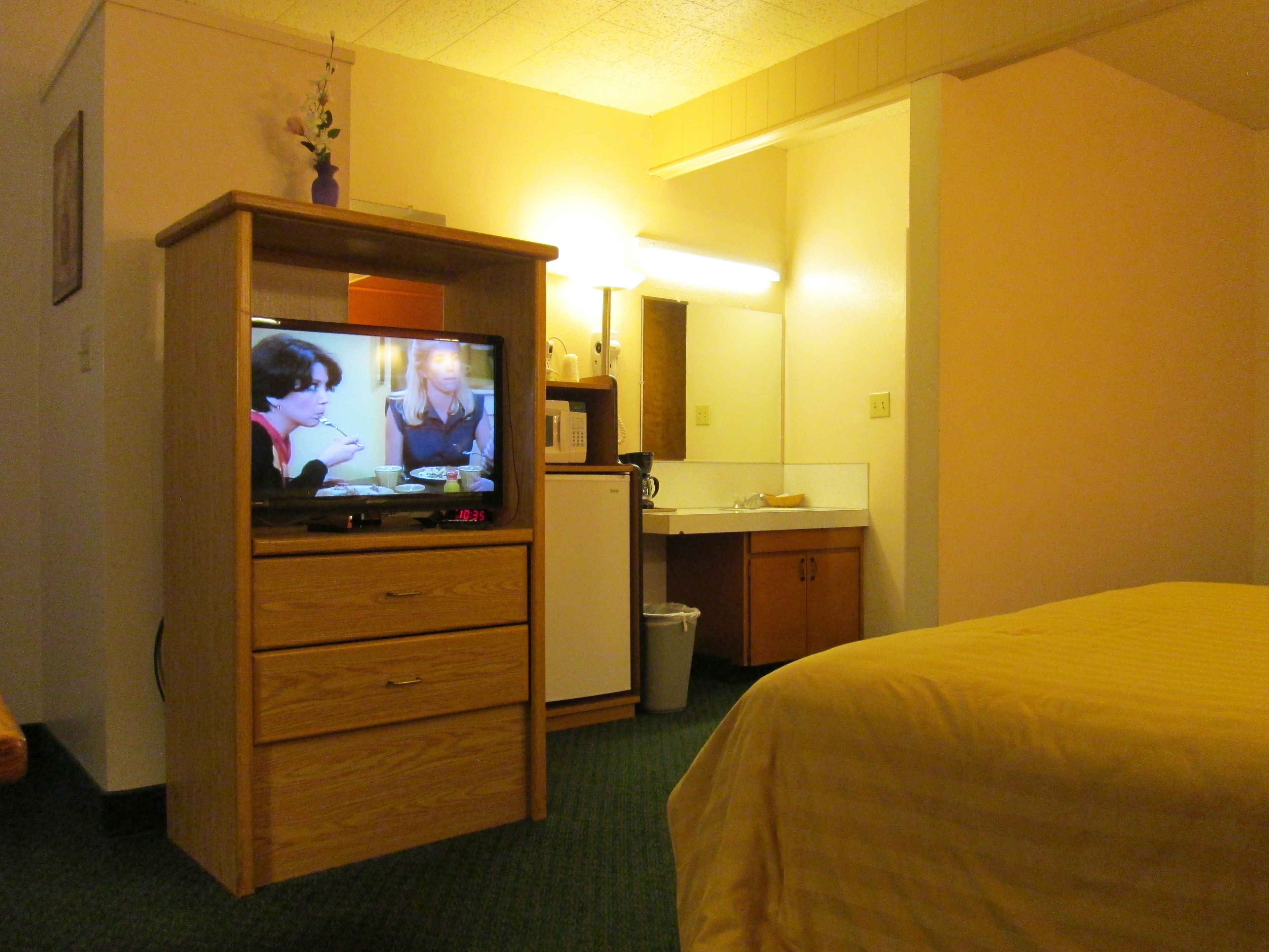 Quarto de hotel com televisão sobre um armário de madeira, um frigorífico pequeno, uma pia e uma cama.
