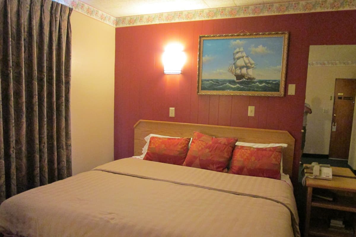 A hotel room with a bed, a nightstand, a painting of a ship on the wall, and a door in the background.