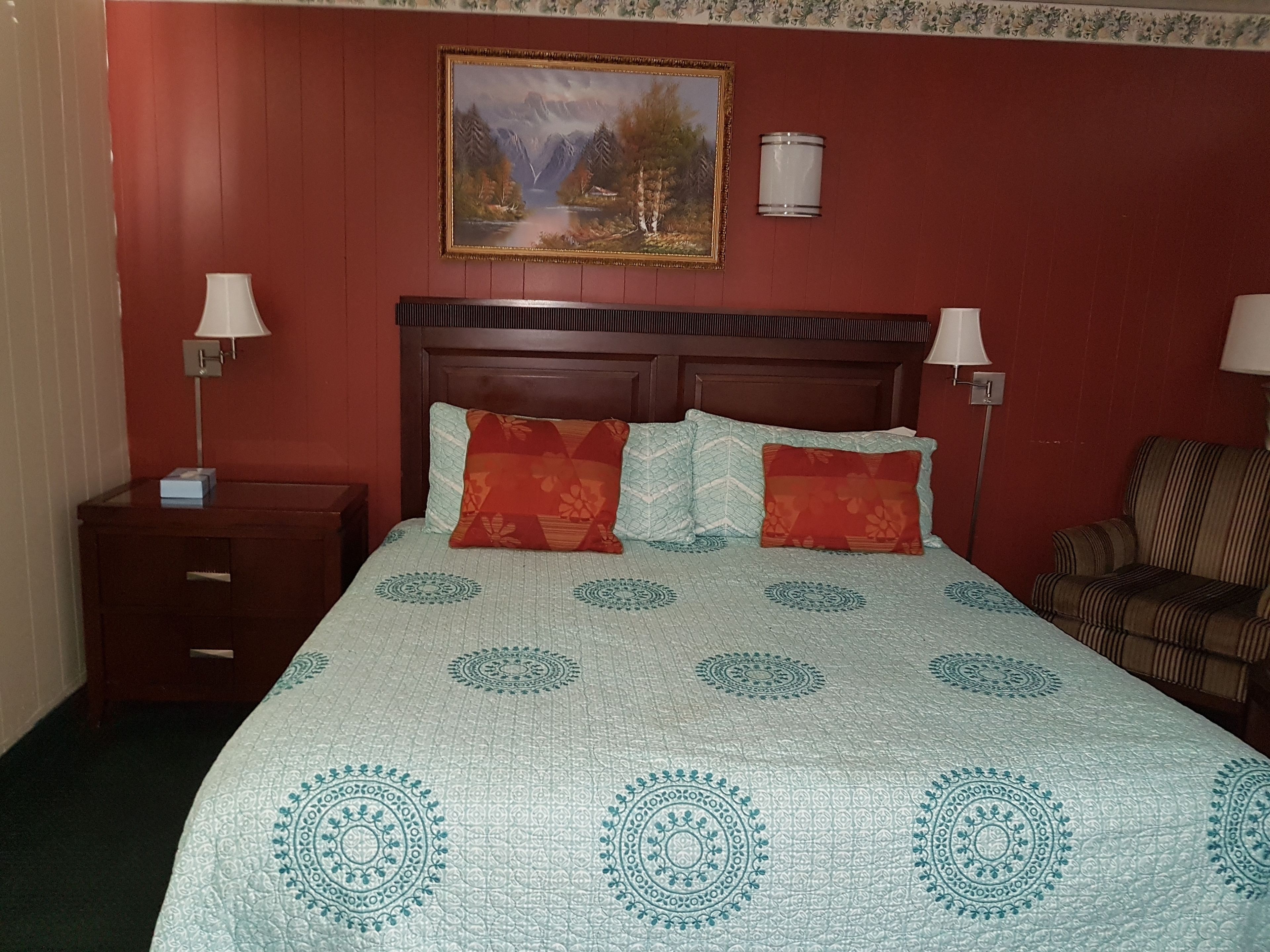 A hotel room with a bed, two bedside tables, a chair, a painting, and a wall-mounted lamp.