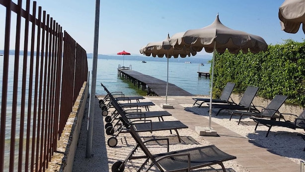 A beachside area with lounge chairs, umbrellas, and a wooden pier extending into the water. - Alexandra Stay Residence (Sirmione)