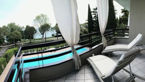 A balcony with a pool, white cushioned chairs, and a view of trees and a body of water. - Alexandra Stay Residence (Sirmione)