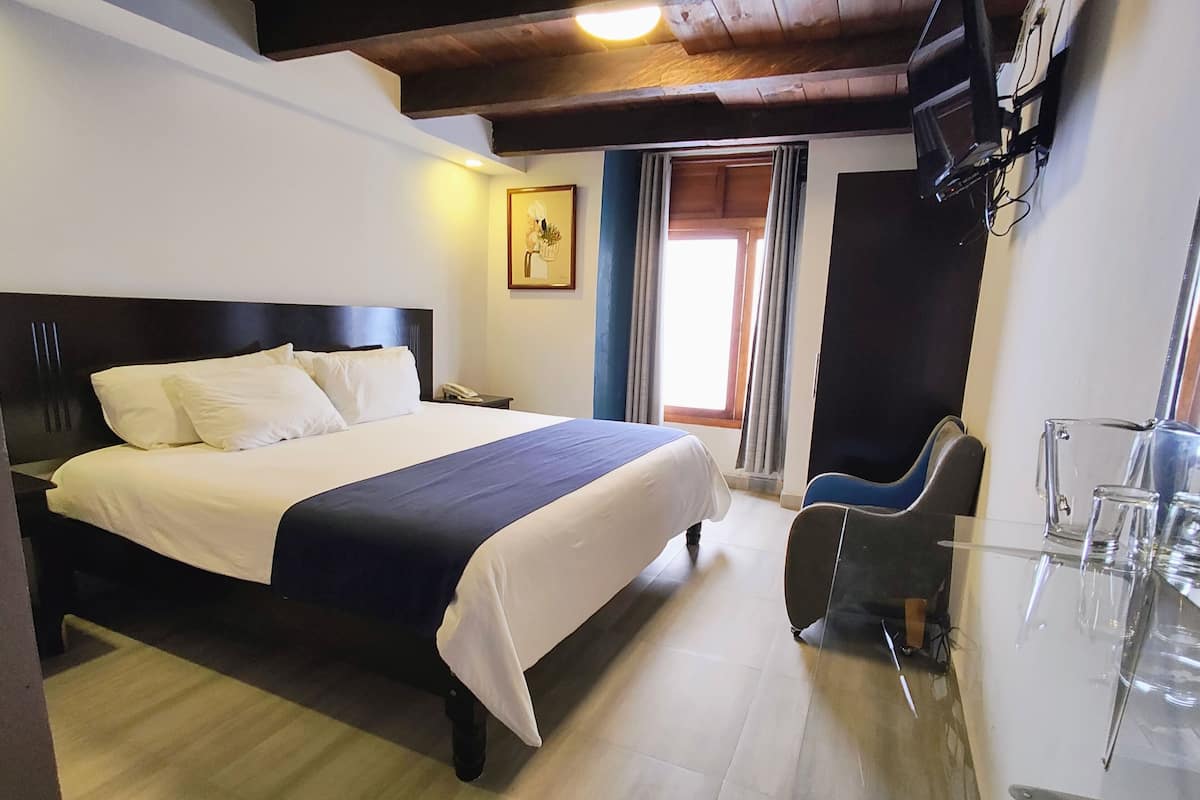 Ein Hotelzimmer mit einem großen Bett, einem Schreibtisch mit Stuhl, einem an der Wand montierten Fernseher und einem Fenster mit Vorhängen.