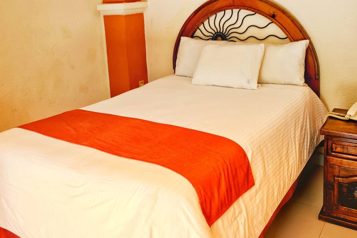 Ein Bett mit weiß-orange gestreifter Bettdecke, ein Holzkopfbrett mit Sonnenelement und ein Nachttisch mit Telefon.