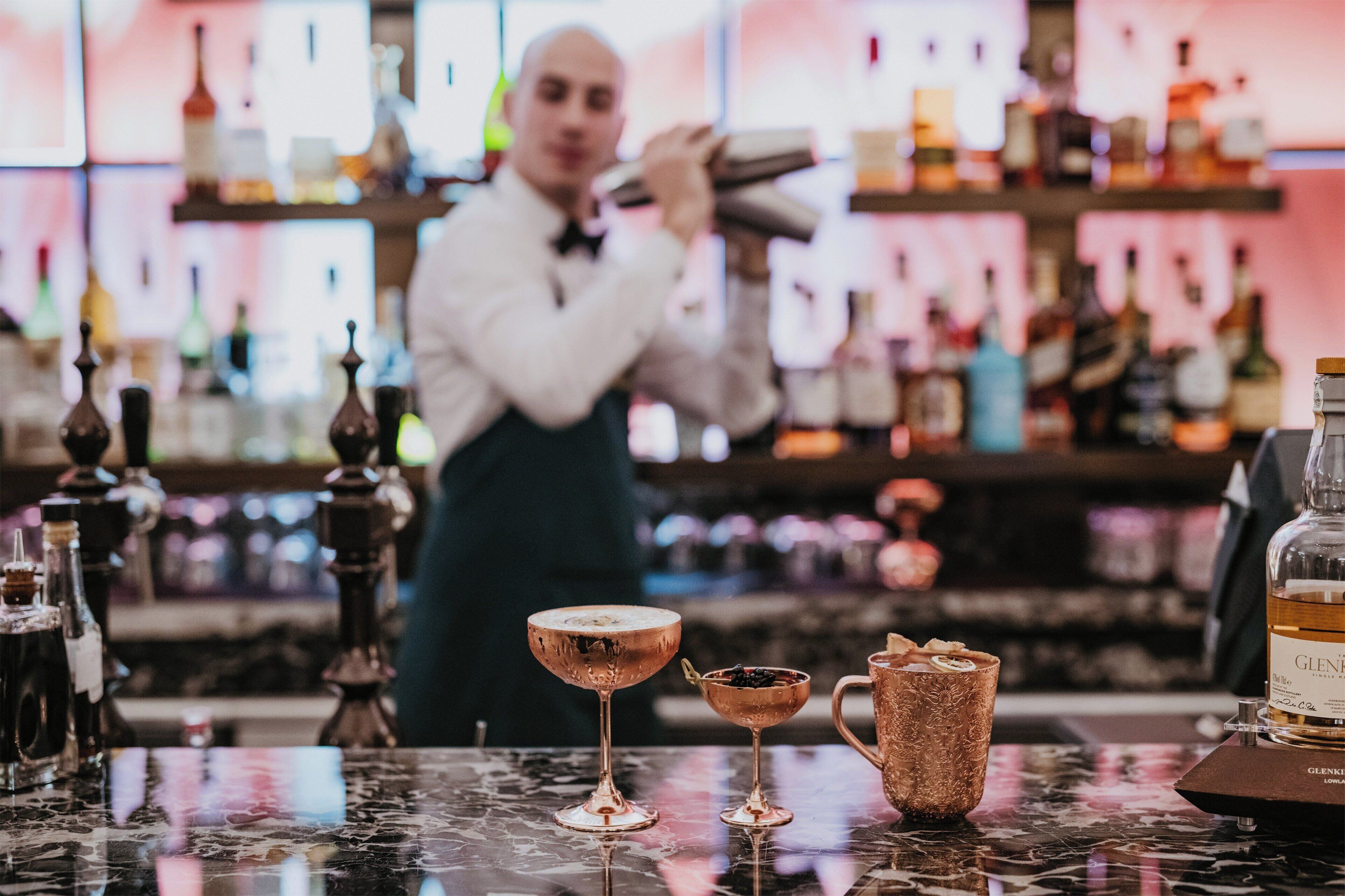 Uma área de bar com um bartender preparando bebidas, diversos frascos e alguns copos no balcão.