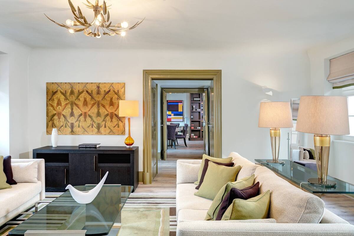 A modern living room with a glass coffee table, a sofa, and two lamps. There is a bookshelf and a decorative wall piece in the background.