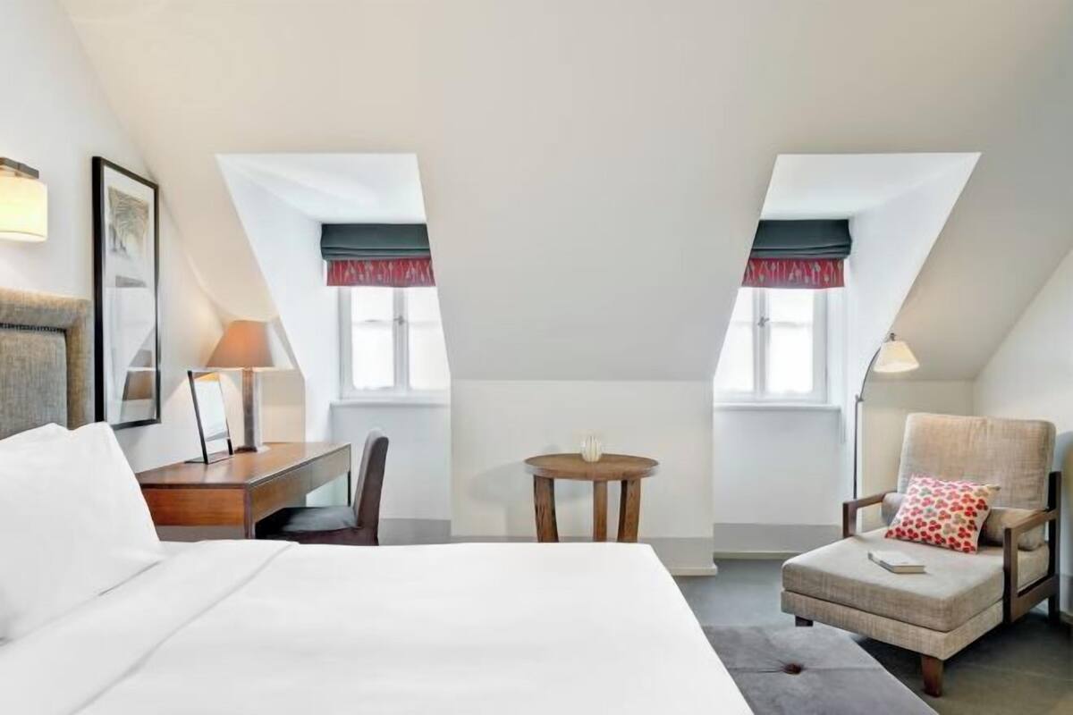 Una habitación de hotel con cama, escritorio, silla y reposapiés. Hay dos ventanas con cortinas.