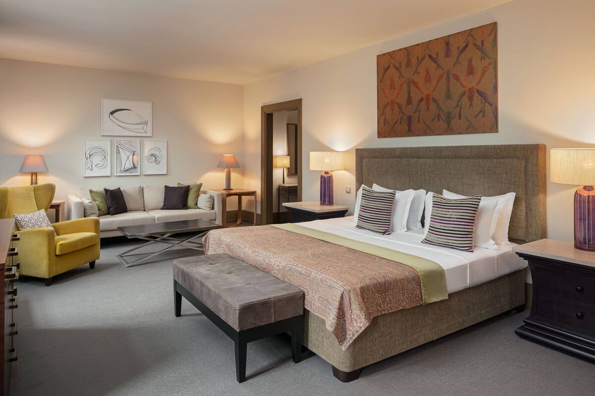 A hotel room with a large bed, a sofa, a chair, a small table, and a nightstand.