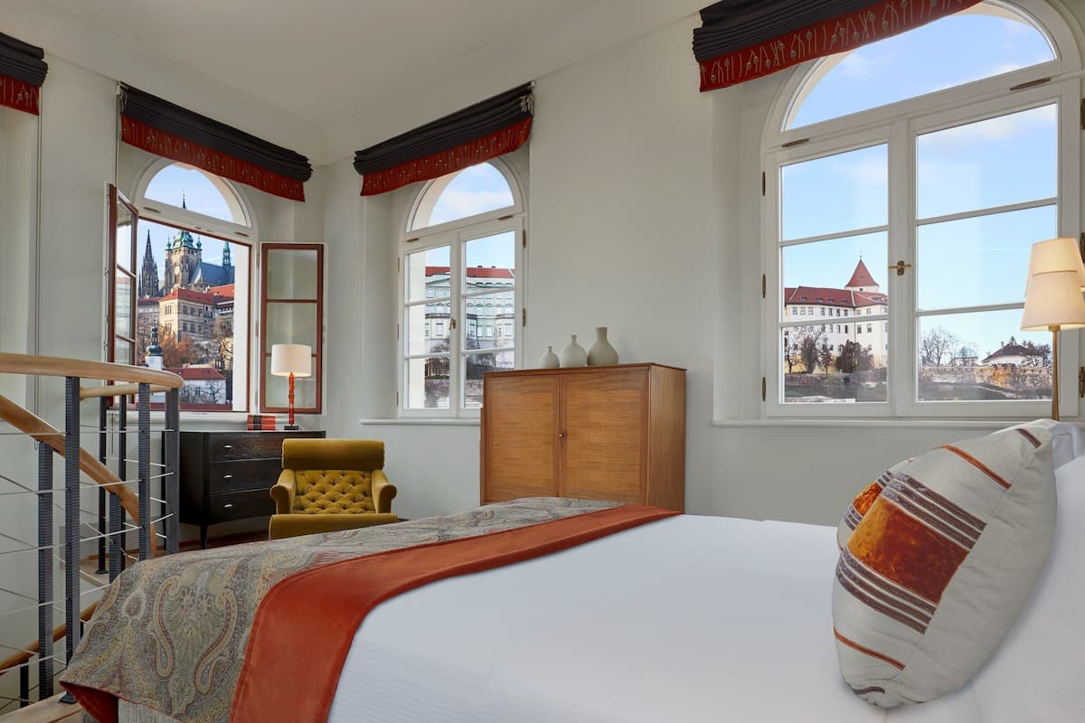 A hotel room with a bed, a chair, a wooden cabinet, and a view of a cityscape through large arched windows.
