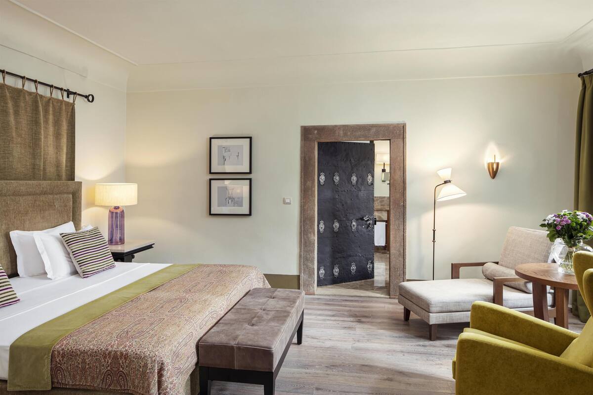 A hotel room with a bed, bedside table, lamp, chair, and a view through an open door.