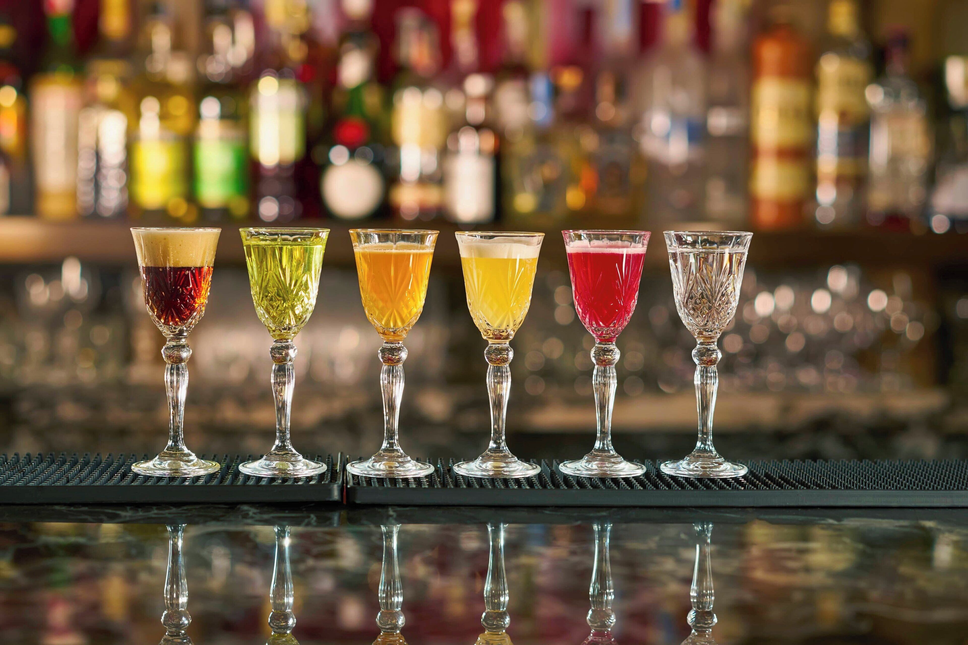 Um balcão com cinco copos de bebidas de cores variadas sobre uma superfície refletora.