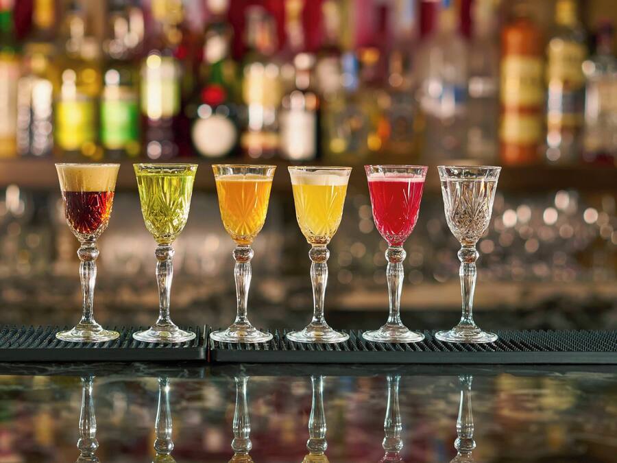 Um balcão com cinco copos de bebidas de cores variadas sobre uma superfície refletora.