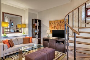 A modern living room with a sofa, ottoman, glass coffee table, and a TV. There's a bookshelf, a mirror, and a painting on the wall.