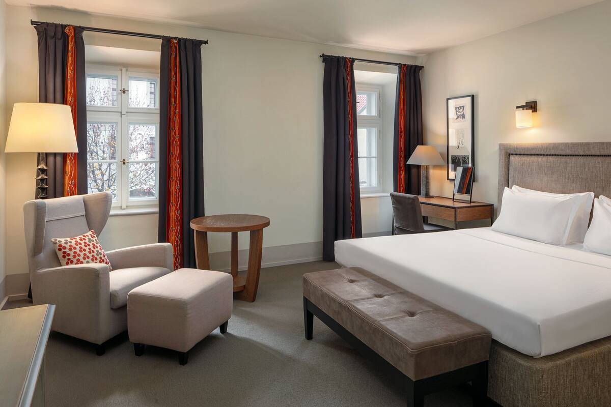 A hotel room with a bed, a chair, a small table, and a bench.
