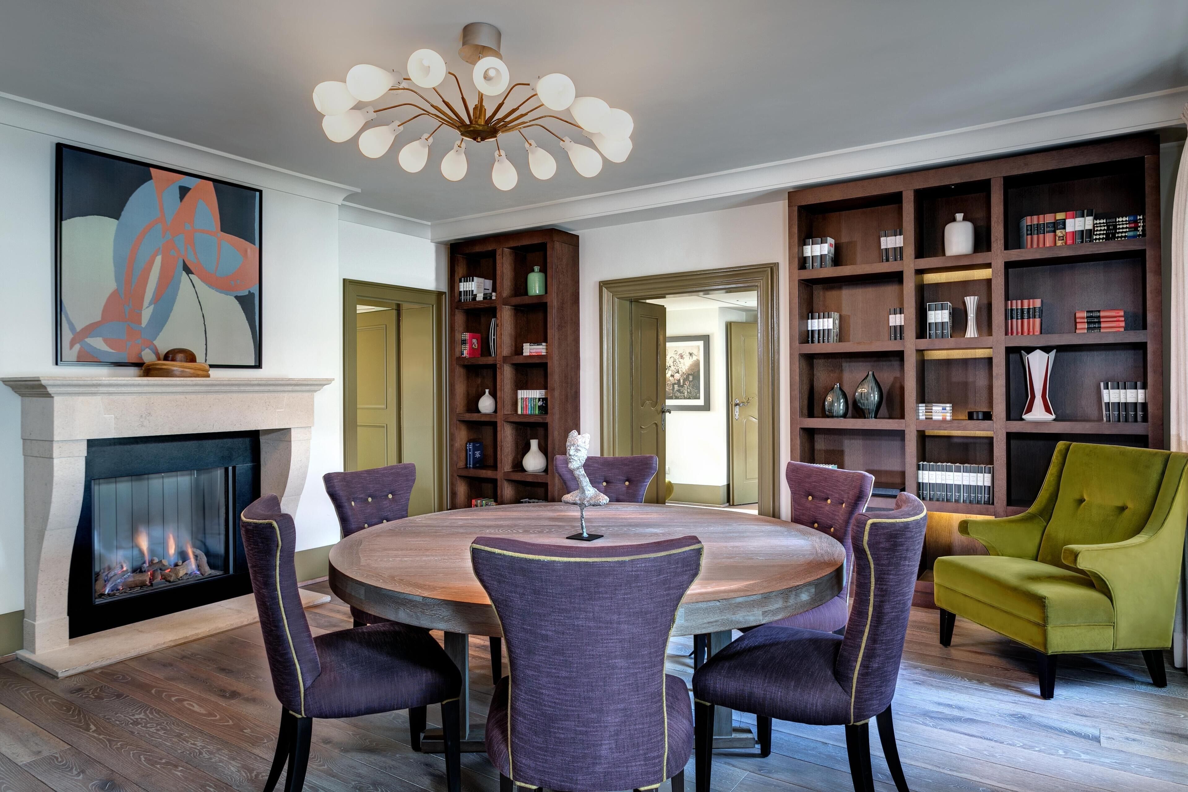 A dining area with a round table, purple chairs, a fireplace, a bookshelf, and a green armchair.