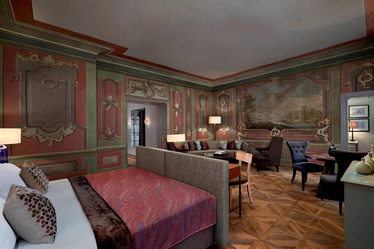 A hotel room with a large bed, a sofa, a dining table, and a chair. There is a painting on the wall and a lamp on the table.