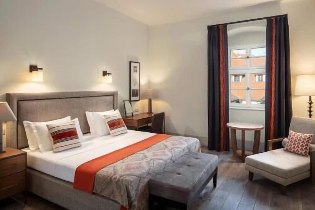 A hotel room with a bed, a chair, a small table, and a window with curtains.