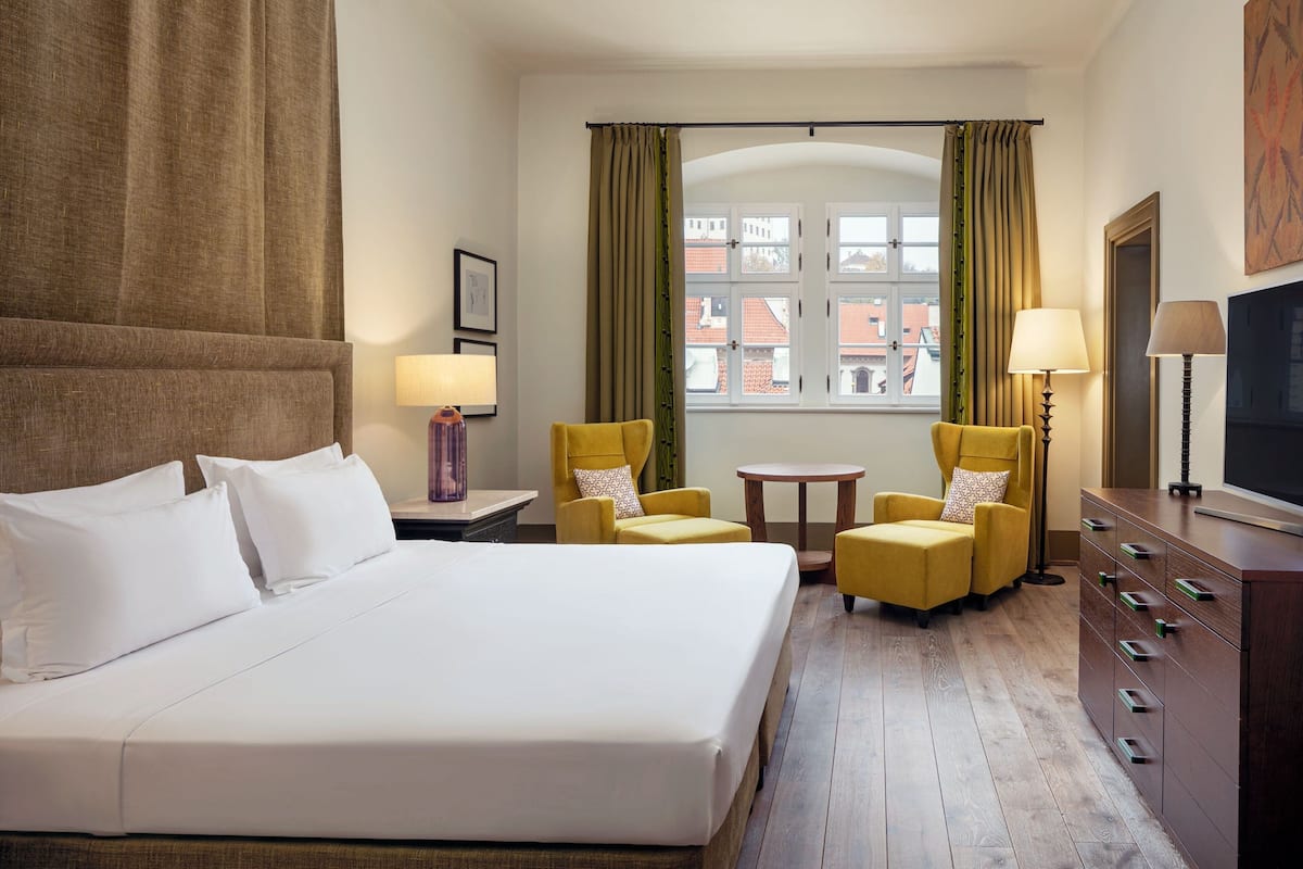 A hotel room with a large bed, two yellow armchairs, a small table, a TV, and a window with curtains.