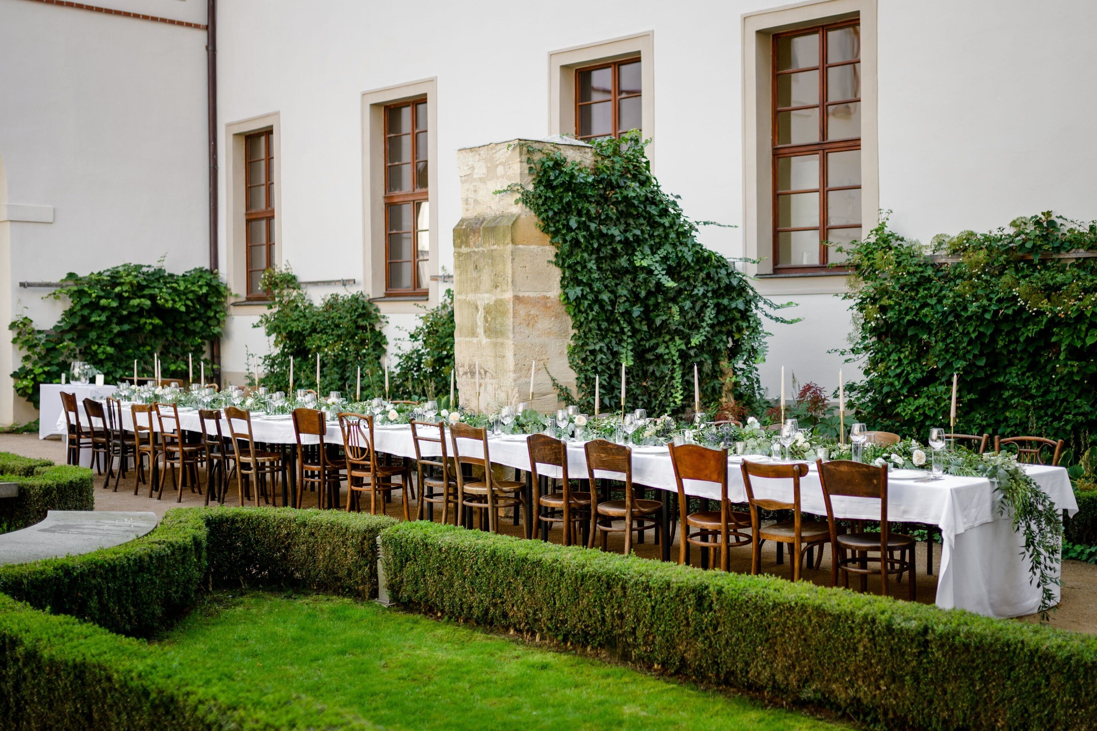 An outdoor dining area with a long table set for a meal, surrounded by chairs and greenery.
