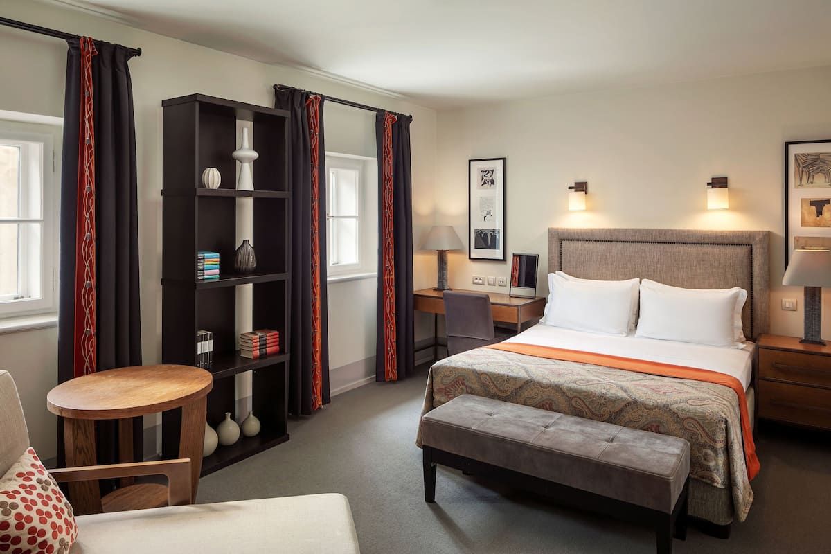 A hotel room with a bed, a desk, a chair, a small table, a bookshelf, and a bench.