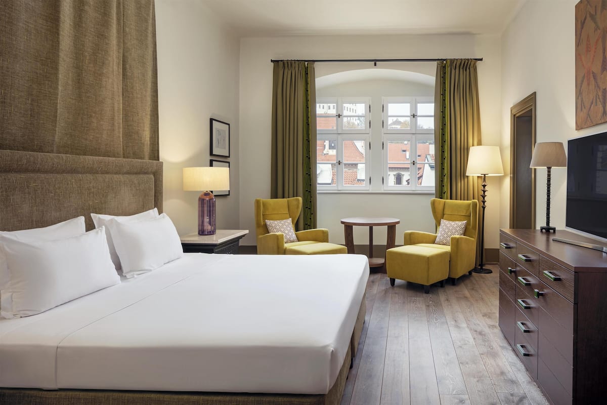 A hotel room with a large bed, two armchairs, a small table, a TV, and a window with curtains.