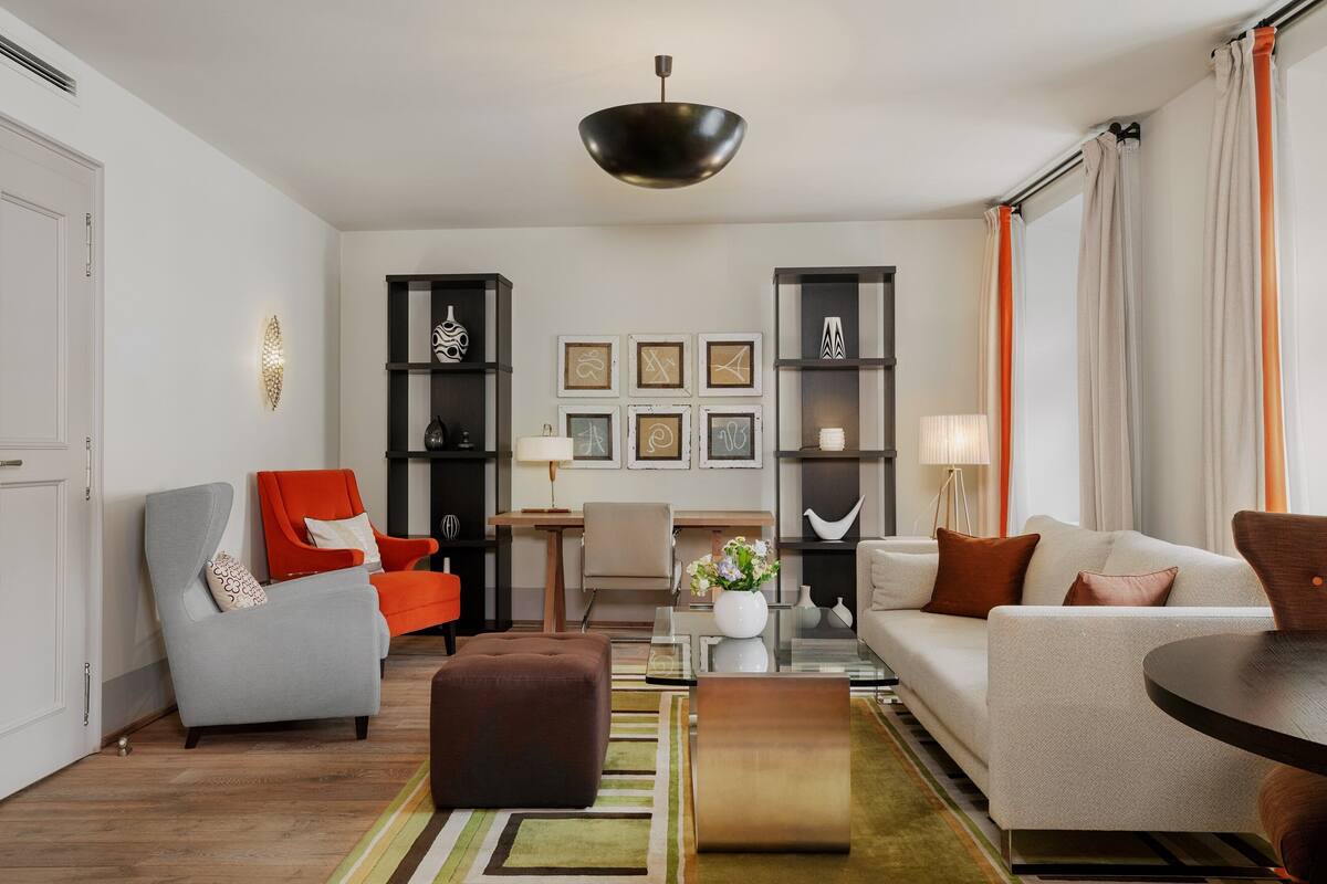 A modern living room with a grey armchair, a red armchair, a white sofa, a brown ottoman, a round coffee table, and a black pendant light.