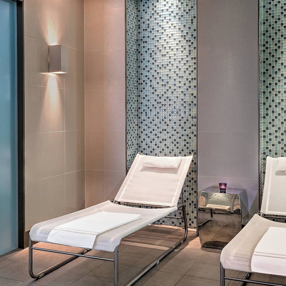 A modern bathroom with two white sun loungers, a tiled wall with a mosaic pattern, and a small table with a candle.