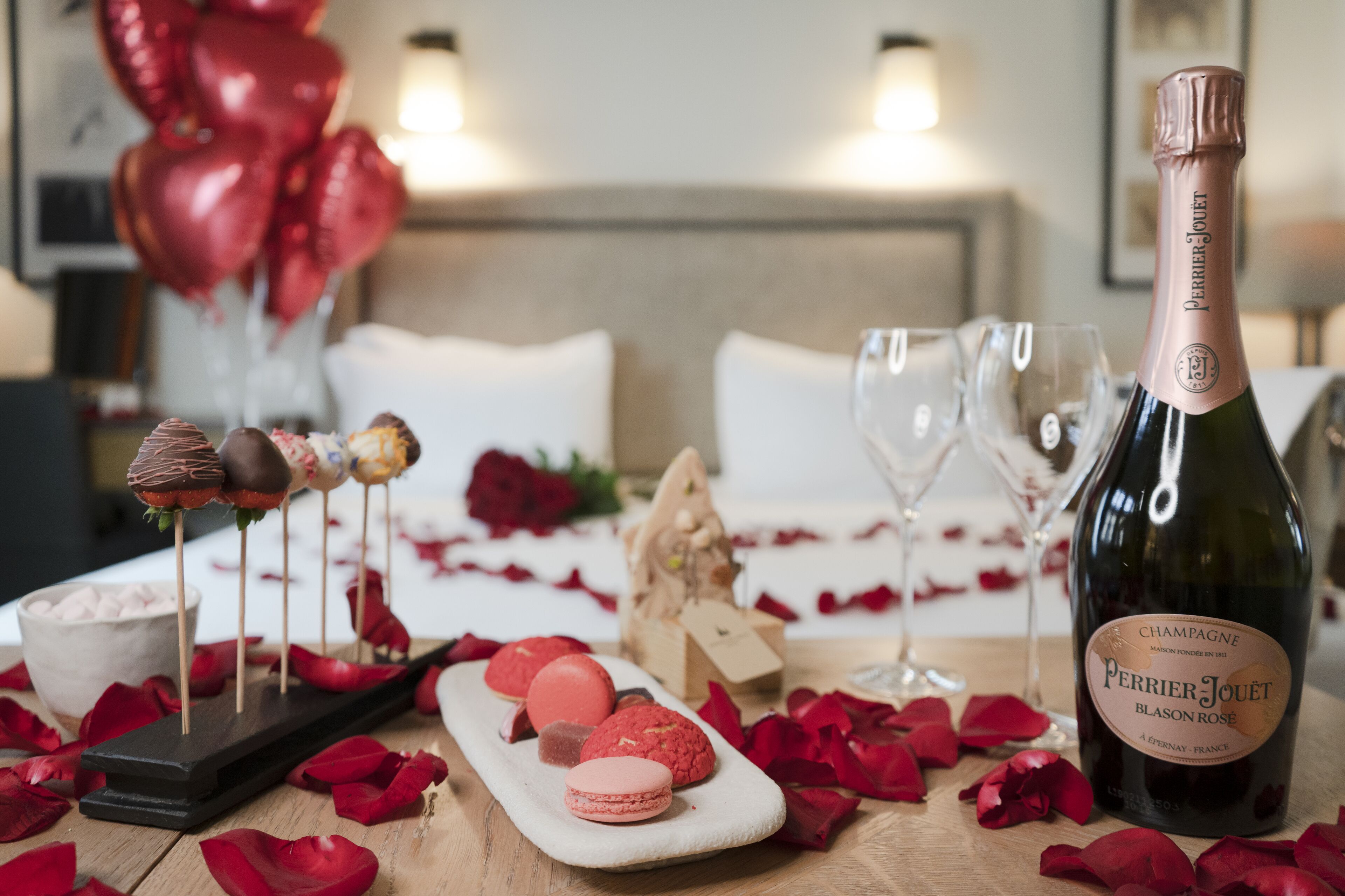 A table set with a bottle of Perrier Jouet Blason Rose champagne, macarons, and chocolate-covered treats on sticks, surrounded by rose petals.