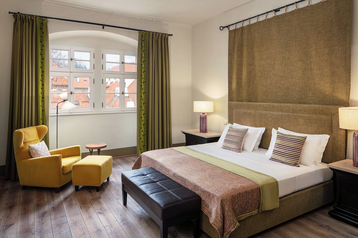 A hotel room with a bed, a chair, an ottoman, a nightstand, and a window with curtains.
