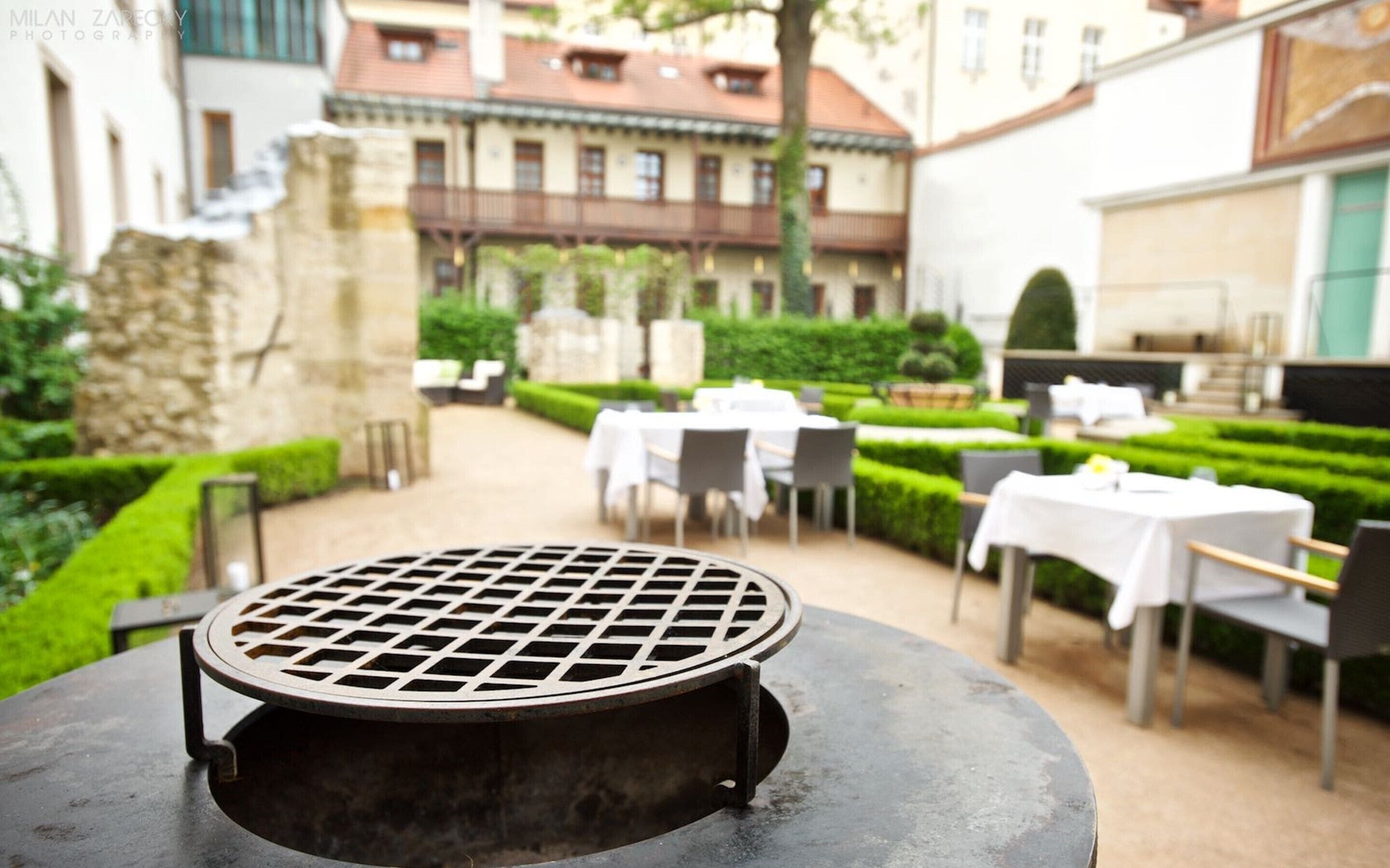 Een terras met een ronde tafel en een barbecue in de voorgrond, omringd door keurig opgestelde witte tafels en stoelen. Achtergrond: gebouwen met balkons en een verzorgde tuin.